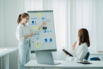 Two young Asian business women meet to analyze the financial chart at the office to discuss the financial situation at the company. A partner sits at a desk with modern documents and equipment.