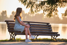 Lonely Woman Sitting Alone On Lake Shore Bench On Warm Summer Evening. Solitude And Relaxing In Nature Concept