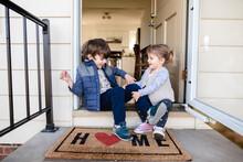 Children Sitting On Front Porch Of Home With Welcome Mat
