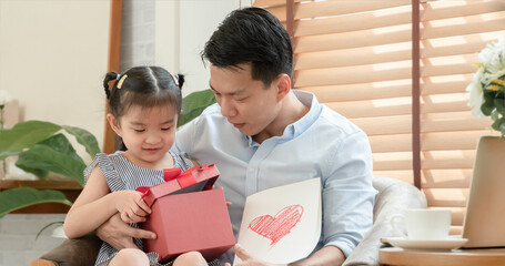 Happy Asian young father surprise with red gift box and heart colored card which little cute daughter give him and sitting on lap while he is working with laptop in living room at home on father's day