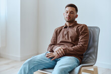Frustrated Sad Young Man Guy Leans On Chair Folded Hands Looks Aside Troubled With Problems Can Not Make Decision Have Mental Psychological Trauma After Bad Break Up Sitting At Home. Copy Space