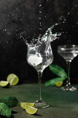 A splash of cocktail in a glass on a dark background with lime slices and leaves