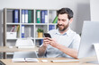 © Liubomir - happy handsome office worker sitting at a computer desk in the office using a mobile phone. Cheerful bearded business man entrepreneur employee. browses smartphone reads messages chats communicates