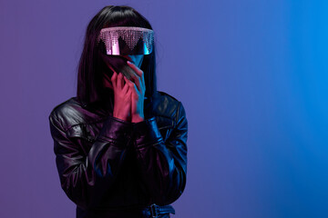Wall Mural - Scared shocked awesome brunet woman in leather jacket trendy specular sunglasses fold hands near face posing isolated in blue violet color light background. Neon party Cyberpunk concept. Copy space
