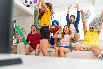 Group of Asian people friends sit on sofa watching and cheering soccer games competition on TV together at home. Happy man and woman sport fans shouting and celebrating sport team victory sports match