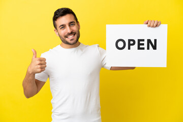 Young handsome caucasian man isolated on yellow background holding a placard with text OPEN with thumb up