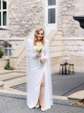Beautiful Blonde Caucasian Bride In A White Dress With A Slit Posing On The Background Of An Old House