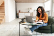 © Prostock-studio - Smiling woman sitting at table, using computer writing in notebook