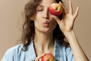 Wall Mural - Funny cute curly female in jeans casual shirt close eye with apple sends kiss at camera posing isolated on over beige pastel background. Healthy food. Natural eco-friendly products concept. Copy space