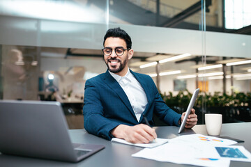 Wall Mural - Happy attractive young islamic male with beard in glasses, suit sits at table with computer and tablet