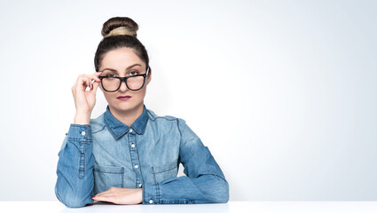 Wall Mural - Serious young woman in a denim jacket, sits at a table, holds glasses with her hand and looks at the camera, on light background.