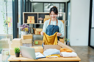 Portrait of a small startup Asian female entrepreneur SME owner picking up a yellow shirt before packing it in an inner box with a customer. Online Business Ideas and Freelance