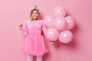 Wall Mural - Festive occasion and holiday concept. Beautiful European woman has worried facial expression wears crown and dress holds magic wand and bunch of inflated balloons isolated over pink background.