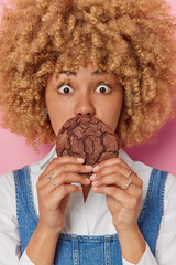 Wall Mural - Vertical shot of shocked curly haired young woman covers mouth with yummy chocolate cookie eats sweet food stunned to break diet dressed in casual clothes poses indoor. Unhealthy food concept