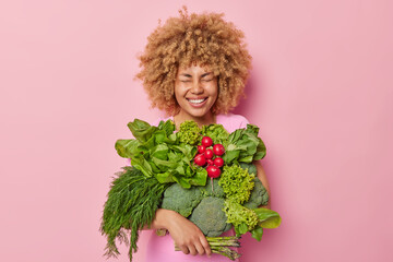 Cheerful woman with curly hair carries different fresh vegetables keeps eyes closed smiles broadly isolated over pink background. Healthy organic food and spring harvest concept. Garden produce