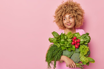 Wall Mural - Clean eating and spring vitamins concept. Satisfied woman with curly hair carries fresh vegetables growned in own garden looks away isolated over pink background blank space for your promotion