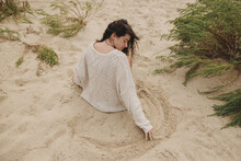 Beautiful Woman With Windy Hair Sitting On Sandy Beach On Background Of Green Grass And Sea, Carefree Tranquil Moment. Stylish Young Female In Knitted Sweater Holding Sand And Relaxing On Coast