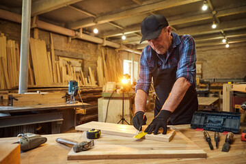 Wall Mural - Portrait of a senior carpenter working with a wood, marking plank with a pencil and taking measurements to cut a piece of wood to make a piece of furniture in the carpentry workshop