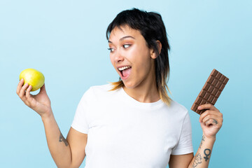 Wall Mural - Young Uruguayan woman over isolated blue background taking a chocolate tablet in one hand and an apple in the other