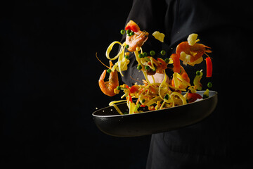 Wall Mural - A bright festive dish - a mix of seafood, Italian pasta and vegetables in a frying pan. Explosion of colors and colors. The process of cooking seafood by a professional chef on a black background.