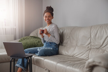 Wall Mural - young beautiful mixed-race female sitting on couch and smiling to camera holding cup of tea