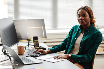 Warm toned portrait of black young woman as IT developer or QA engineer smiling at camera while sitting at workplace with computers, copy space