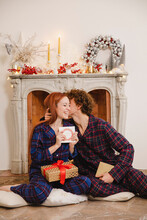 Man Kissing Girlfriend Holding Greeting Card Sitting With Gift In Front Of Fireplace At Home
