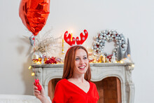 Smiling Woman Wearing Christmas Headdress Sitting With Red Heart Shape Balloon At Home