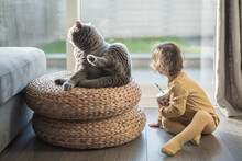 A Toddler Wearing Yellow Pyjamas And Holding A Cup Looks Back Mimicking Her British Short Hair Cat Sitting On A Wicker Stool In A House In Edinburgh, Scotland, UK