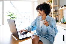 Smiling Woman Holding Coffee Cup Using Laptop Sitting At Kitchen Island