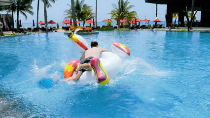 Wall Mural - Happy funny man successfully jump onto floating inflatable unicorn in swimming pool in tropical hotel to ride. Crazy male tourist have fun on giant toy in water. Summer vacation, holiday concept.