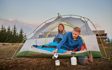 Wall Mural - Husband, after waking up in the morning, lying in white tent, pouring freshly brewed hot coffee into metal cup for his wife. Active rest in camp in the mountains. Concept of hiking and camping.