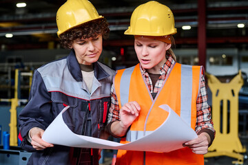 Two young female engineers discussing sketch on blueprint at working meeting at factory while one of them commenting its details