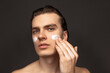 Facial skin care for man. Young happy man with perfect well-kept skin using face cream isolated over grey background. Cosmetics, health care, skin care, beauty