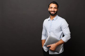 Cheerful Indian bearded man holding laptop standing isolated on black, looking at camera with happy smile. Male employee or freelancer carrying laptop computer. Copy space