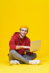 Happy young hipster man in red hoodie sitting on the floor holding laptop studying or working. Smart freelancer caucasian man with laptop isolated on yellow background