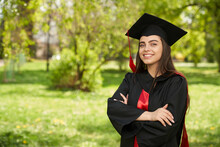 Front View Of Girl Wearing Mortarboard And Graduate Gown Smiling. Pretty Young Female Graduating From College, Standing With Crossed Hands, Looking At Camera. Concept Of Youth.