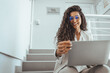© Dragana Gordic - Focused young business woman working on a laptop on some stairs on hallway. Young businesswoman using laptop on steps. Cropped frame of smiling female professional