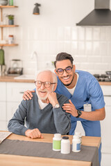 Wall Mural - Cheerful caregiver embracing a pensioner seated at the kitchen table