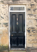 Traditional Old Black Painted Wooden House Door With Brass Letter Box And Knocker With A White Frame Surrounded By A Stone Wall
