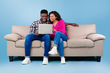 Wall Mural - Happy young black couple using laptop while sitting on sofa over blue studio background, full length