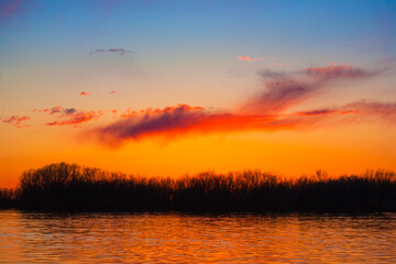 Orange, yellow and purple sunset on river with dark colorful clouds in sky with trees reflection in water