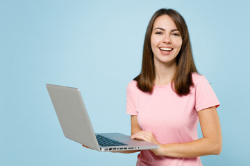 Wall Mural - Young smiling freelancer cool happy caucasian woman 20s in pink t-shirt hold use work on laptop pc computer isolated on pastel plain light blue background studio portrait. People lifestyle concept