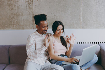 Wall Mural - Two young friends woman man of African American ethnicity in casual clothes hold use laptop pc computer talk by video call waving hand sit on gray sofa indoor spend free time rest in flat together.