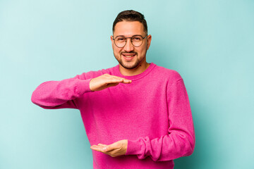 Wall Mural - Young hispanic man isolated on blue background holding something with both hands, product presentation.