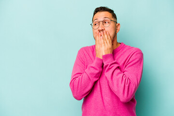 Wall Mural - Young hispanic man isolated on blue background thoughtful looking to a copy space covering mouth with hand.