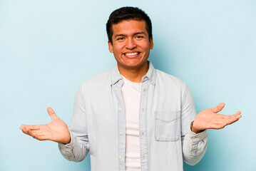 Young hispanic man isolated on blue background makes scale with arms, feels happy and confident.