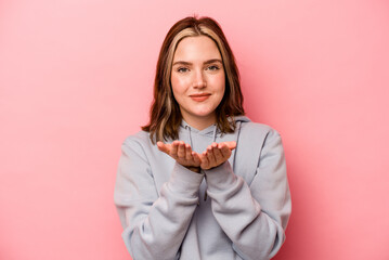 Wall Mural - Young caucasian woman isolated on pink background holding something with palms, offering to camera.