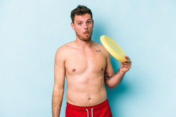 Young caucasian man playing with frisky isolated on blue background shrugs shoulders and open eyes confused.