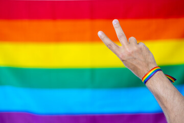 Mano haciendo el símbolo de la paz con pulsera de colores del colectivo lgtbi, con la bander lgtbi de fondo